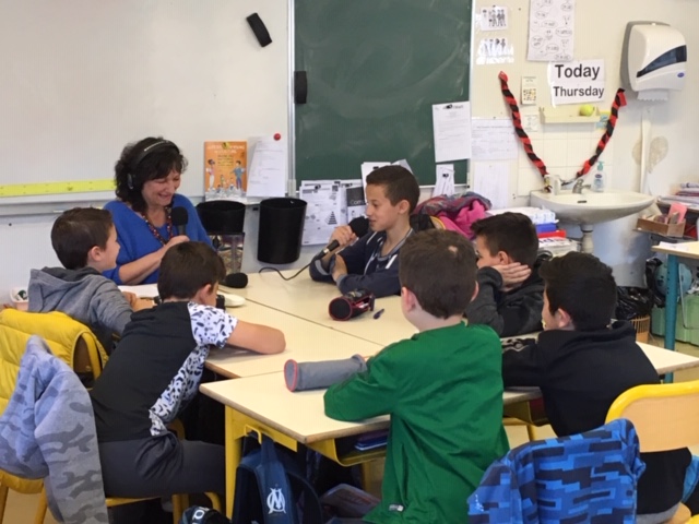 Dans une salle de classe. Autour d'une table, casque sur les oreilles, Sylvie, animatrice radio BLV, entourée de 6 jeunes garçons qui l'écoutent attentivement. Un des enfants parle au micro.